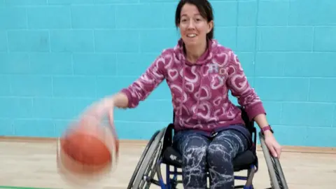 Sally Millington Seorang wanita dengan hoodie ungu dan legging biru memantul bola basket dari kursi roda di ruang olahraga.