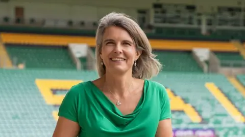 Northampton Saints Julia Chapman, who has shoulder-length blonde hair and is wearing teardrop-shaped earrings, and a green top, stands inside Frankin's Gardens stadium at Northampton Saints. She has a wide green.