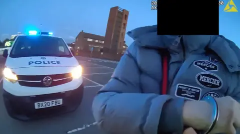 A man with a black box over his face in handcuffs. He is mixed race and wearing a grey Mercier coat with a Gucci belt. There is a police car behind him. 