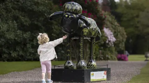 David Oates Photography A young girl is reaching up towards a model of a sheep. She has short, curly blonde hair and is wearing a patterned jacket and pink trousers. The sheep is painted black with gold patterns on its body and legs