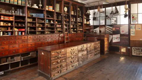 Historic England Archive Interior general view showing shop counter and wooden shelving. There are lots of small mahogany looking drawers with nobs on and various ornaments and glasses tucked into shelves above, a wooden ladder stretching upwards. To the right, skates hang from the ceiling. A sign near the bottom of a wooden wall says 'Starex roofing'. The floor is wooden and there is a grid of windows towards the top of the wall to the right.