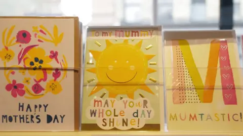 Three Mother's Day cards are pictured on a shop's shelf. One has an image of flowers, another the sun, and a large 'M' on the final card.