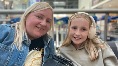 Janine and her daughter sat in the departure lounge of the airport. They both have blonde hair. Janine is wearing a blue denim jacket, and Abbie is wearing a cream hoodie and cream headphones. They are both smiling and looking at the camera