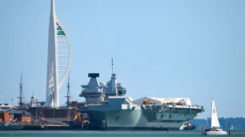 The Royal Navy's HMS Queen Elizabeth aircraft carrier with Portsmouth's Spinnaker Tower in the background, and a small yacht passing in the foreground