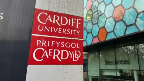 BBC A red sign on a grey plinth reading 'Cardiff University, Prifysgol Caerdydd'. The sign is outside a colourful glass hexagon patterned building with the word biosciences written underneath