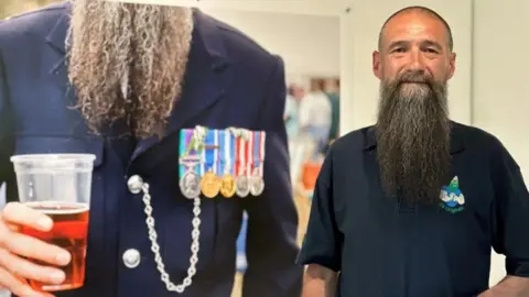 Mark Huston, who has a long beard, standing next to a large photo of himself holding a plastic pint glass of beer while in his prison officer's uniform.