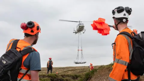 National Trust/Paul Harris A helicopter carries white bags suspended on a line, marshalled by four people on the ground in hi-vis