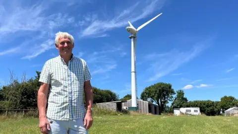 Peter Bailey wearing a checked shirt with the wind turbine and wooden outbuildings and a caravan in the background.