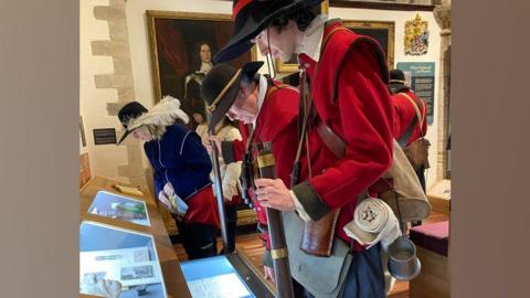 Four men dressed as Civil War characters, three in scarlet coats and a fourth in a blue coat, browse displays in a museum