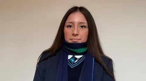 Sophia. She has long dark hair, wearing a green and navy scarf and a navy school uniform.