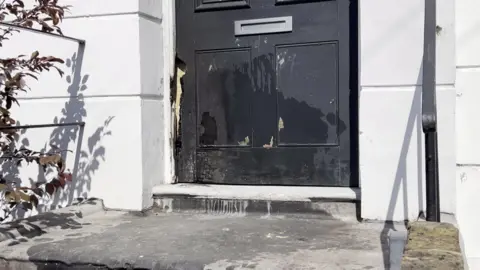 Arrest over suspected arson on properties linked to PM 8 Fire damage on a black door, set in a white building