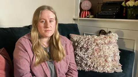 A woman in a pink shirt with long blonde hair sits on a dark green sofa. There are cushions and a blanket next to her. Behind her you can see a typewriter and some roses on a shelf.