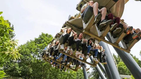 Getty Images A group of visitors are on a ride sitting with their feet dangling from the ride. There are trees in the background and some of the people are smiling. The picture is taken from the ground looking up towards the people.
