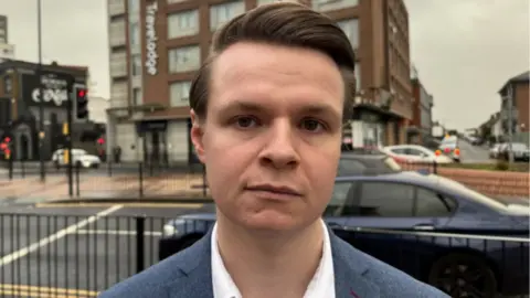 A man wearing a white shirt and a blue blazer stood in front of a main road with a Travelodge in the background. He has a serious expression and is looking straight into the camera.