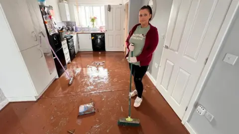 Laurie Price is standing in a flooded kitchen with a brush. The floor is completely brown with dirt. She is wearing a red jacket and a green t-shirt. She is in casual clothing because she is trying to clean the damage. 