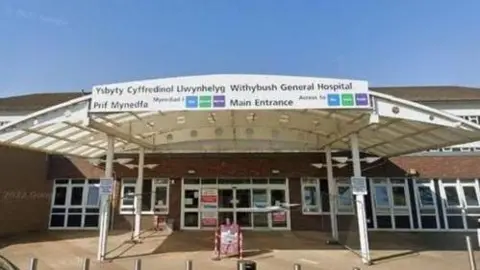 An entrance to a hospital, with a sign at the top and a car parked out front