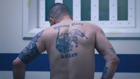 BBC/Expectation Entertainment A tattoo saying "Natural Born Killer" around a design of a dragon-like creature is on the back of a man.