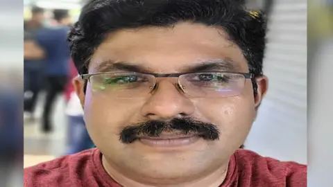 A close-up photograph of Deepak U facing the camera. He has short, dark hair, wears rectangular glasses, and has a thick moustache. His expression is neutral. He is wearing a dark red, short-sleeved T-shirt. The photograph appears to have been taken indoors. Behind him, the background is blurred, with indistinct figures of people and what look like closed shop shutters or metal doors, suggesting a public place