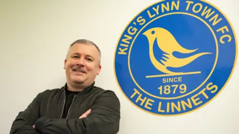 Paul Caddis wearing a black jacket over a black T-shirt. He has his arms crossed and is smiling while looking into the camera. He is standing in front of a white wall on which there is a large blue and yellow King's Lynn Town FC crest. 