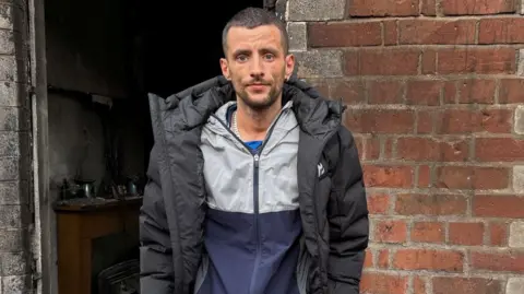 Jamie Corry stands outside of his burnt out home in east Belfast. He is looking at the camera with a neutral expression on his face. He is wearing a black puffed jacket.