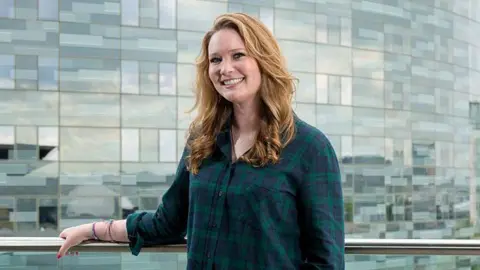 Getty Images A woman with red hair wears a green and navy checked shirt and stands next to the metal railing of a glass balcony overlooking a modern high-rise building.