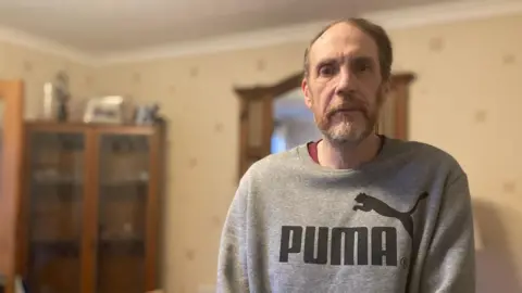 Gary Punter is wearing a grey puma sweatshirt and is standing in the back room of his rental property. He is looking at the camera and has beard. Behind him can be seen a corner, glass-fronted cupboard.
