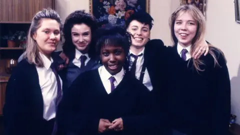 The five actors, dressed in their character's school uniforms, smile at the camera