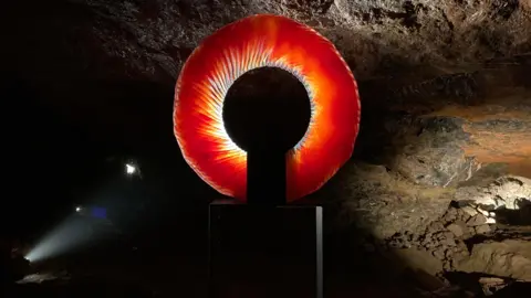 A sculpture made of glass sits inside a cave. It is a red hoop that partly lets light through, with what looks like lines carved into it.