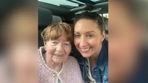 Darren Mullen Rachel Duffy and her mother Anne are sitting in a car. They both have their heads against one another and are smiling. Anne is an elderly woman with short brown hair. She is wearing a pink and white jumper with a gold button. Rachel has her brown hair tied back in a bun. She has a pair of sunglasses on her head and is wearing a denim jacket.