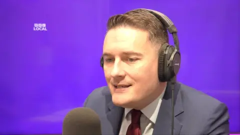 The health secretary in a radio studio with a red tie and wearing headphones. The background is purple.
