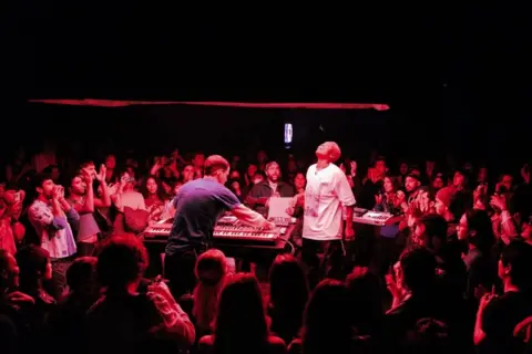 Arlo Parks Arlo Parks and her producer Baird play on a small stage, surrounded by fans, in a small Los Angeles venue