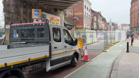 The entrance to a road under a bridge has been cordoned off. There is a parked white van and metal fencing and an orange and white cones which blocks off the entrance. In the background are shops and people.