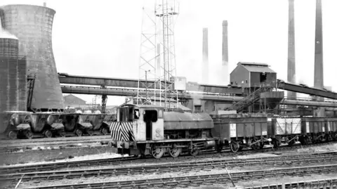 NLR A black and white image of a steam locomotive at Corby steel works. The train is on a railway track, with steam coming out of it, with buildings behind it, mostly chimneys.