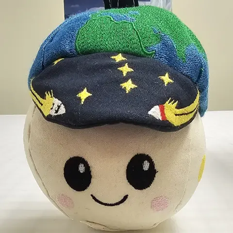 NASA A small, round soft toy sits on a white tabletop, about the size of a large grapefruit. Its fabric face is pale cream with big black embroidered eyes, rosy pink cheeks and a simple smiling mouth, giving it a cute cartoon look. On top, it wears a dark blue cap sprinkled with embroidered yellow stars and tiny white rockets. Above the cap is a padded green‑and‑blue globe, like a miniature Earth, with a short dark loop for hanging. The toy’s overall impression is friendly and playful, like a cheerful mascot for a children’s space adventure.
