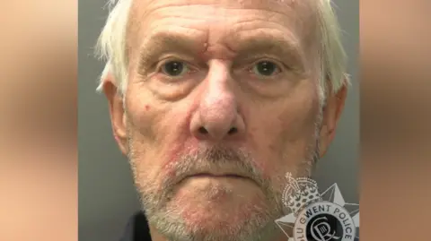 A police custody image of Peter Rowell. He is in his 60s with messy grey hair and short grey stubble. The collar of a dark shirt is visible. Gwent Police's Logo sits in the bottom right-hand corner. 