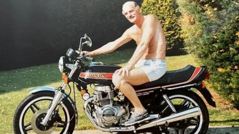 Sally Oliver The picture shows a person sitting on a classic Honda motorcycle. The bike has a black frame with red and orange striping and chrome detailing on the exhaust and engine. The setting is an outdoor garden or yard with green grass and tall shrubs in the background. The person is wearing light shorts and trainers, and the motorcycle is parked on a paved surface. 