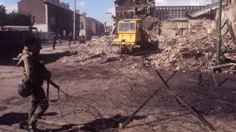 PA Media A still from 1969 of a soldier at a barbed wire barrier as there is a digger clearing up rubble on the Falls Road in Belfast.