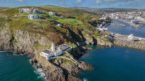 An arial view of Douglas Head, you can see the lighthouse on a rocky terrain, there are buildings scattered on the green terrain.