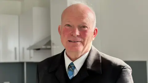 Christopher Brown, who is balding and it wearing a smart black coat over a white shirt and blue Manx tartan tie.