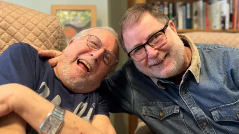 Bernard Mendoza and his son Danny sat down and smiling at the camera. Danny, wearing a blue denim shirt and reading glasses, has his arm around his father, who is also wearing glasses and a blue t-shirt.