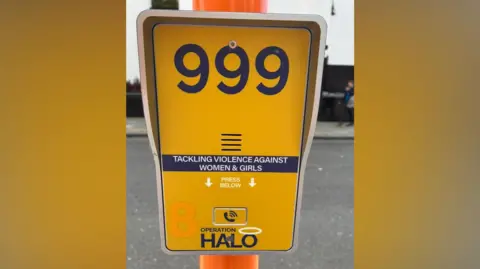 Marc Gaier/BBC A yellow metal intercom with a call button fitted to an orange lamp-post. The numbers 999 are painted onto the box in grey followed by the phrase 'Tackling Violence Against Women & Girls' and then 'Operation Halo'.