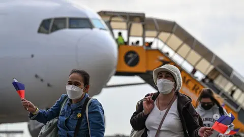 Three women waving the Philippine flag and wearing face masks, walking from an airplane in the background after being repatriated fro the Middle East due to the US and Israel's war on Iran