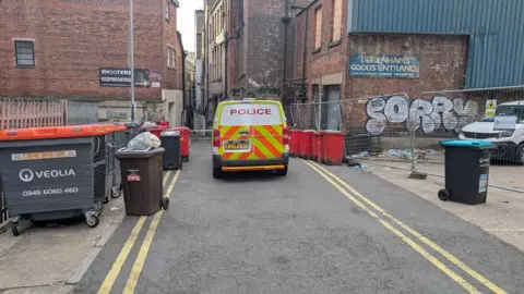 Police van parked by bins at the rear of older, brick shop buildings