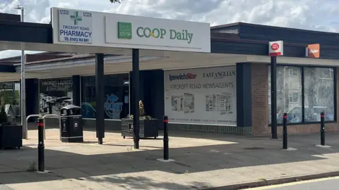A pavement stretching left to right, behind which is a wooden structure covering a paved shopping area with black bins and planters, flanked by shops. A sign above the entrance says Co-Op Daily and Fircroft Road Pharmacy.  