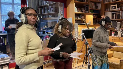 Lily-May Symonds/BBC Three women wearing headphones in front of a microphone on a stand in a recording studio. Two of the women are wearing jumpers and one is wearing a coat. There is a man in the background holding a piece of paper. There are shelves full of records in the background and a leather armchair on the right.
