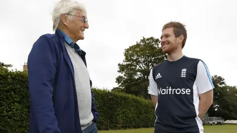 Getty Images  Sir Michael Parkinson, wearing a blue jacket, grey jumper and light blue shirt under it, talking to Eoin Morgan, dressed in an England cricket top, in front of a tree and a hedge at the cricket club 