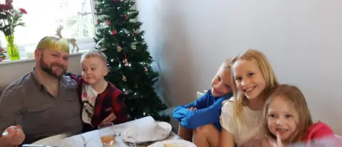 Gemma Cole James sitting with four children around a dinner table around Christmas time. A Christmas tree can be seen in the background. All are smiling.