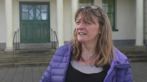 Nikki Hurst has long brown hair. She is wearing a purple coat and a black top. She is standing in front of a grey building with a green door.
