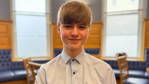Jasper has short light brown hair. He is wearing a light blue shirt buttoned to the top. He is standing in a room panelled in pine wood with blue at the top part of the wall. At the bottom is banquette seating in blue leather and several chairs behind him.