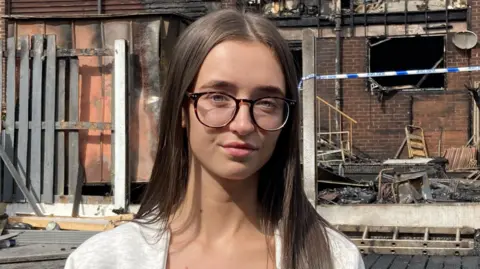 Malik Walton/BBC A young woman with long brown hair and round glasses stands in front of a burned-out garden.
